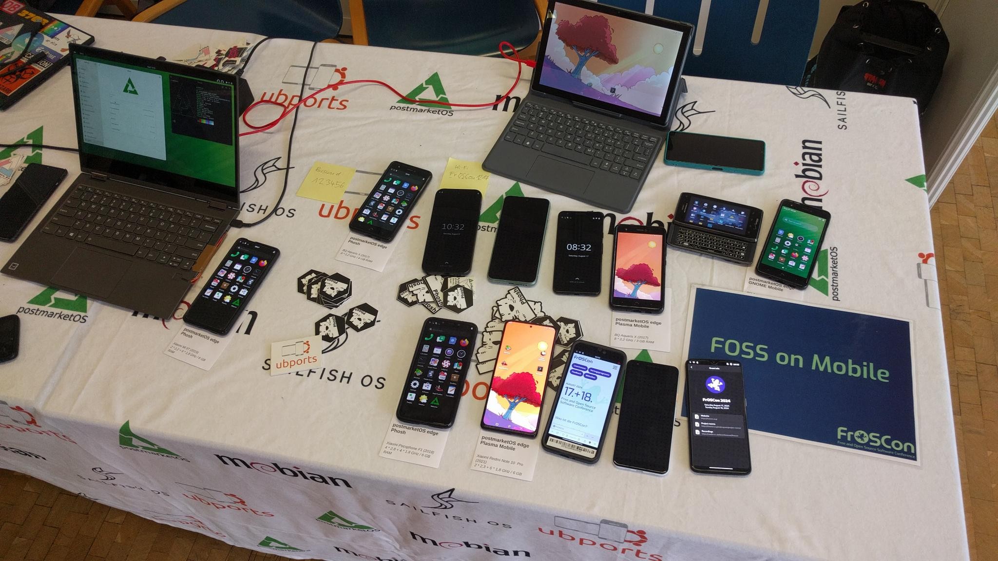 FOSS on Mobile stand at FrOSCon, a table with the FOSS on mobile tablecloth, a laptop, a tablet, a bunch of phones all running various Linux Mobile distributions
